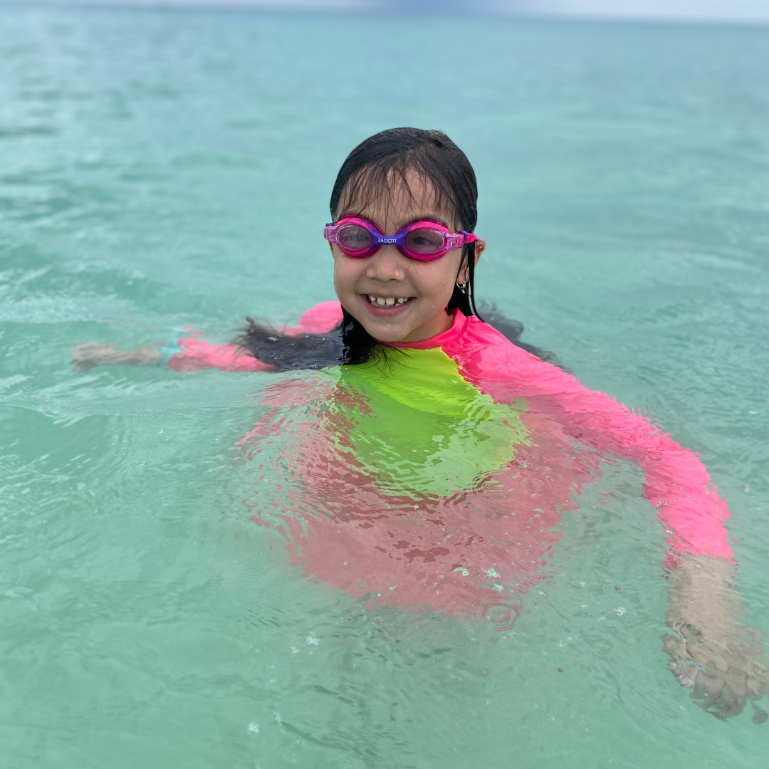 Red haired pre-school boy with high visibility neon rash guard sun shirt pouring water from a green bucket at the beach.  