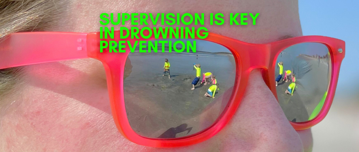 Photo of a woman wearing sunglasses with the reflection of her kids in neon swimwear on the beach in the center focus demonstrating that she is supervising them closely to prevent drowning.