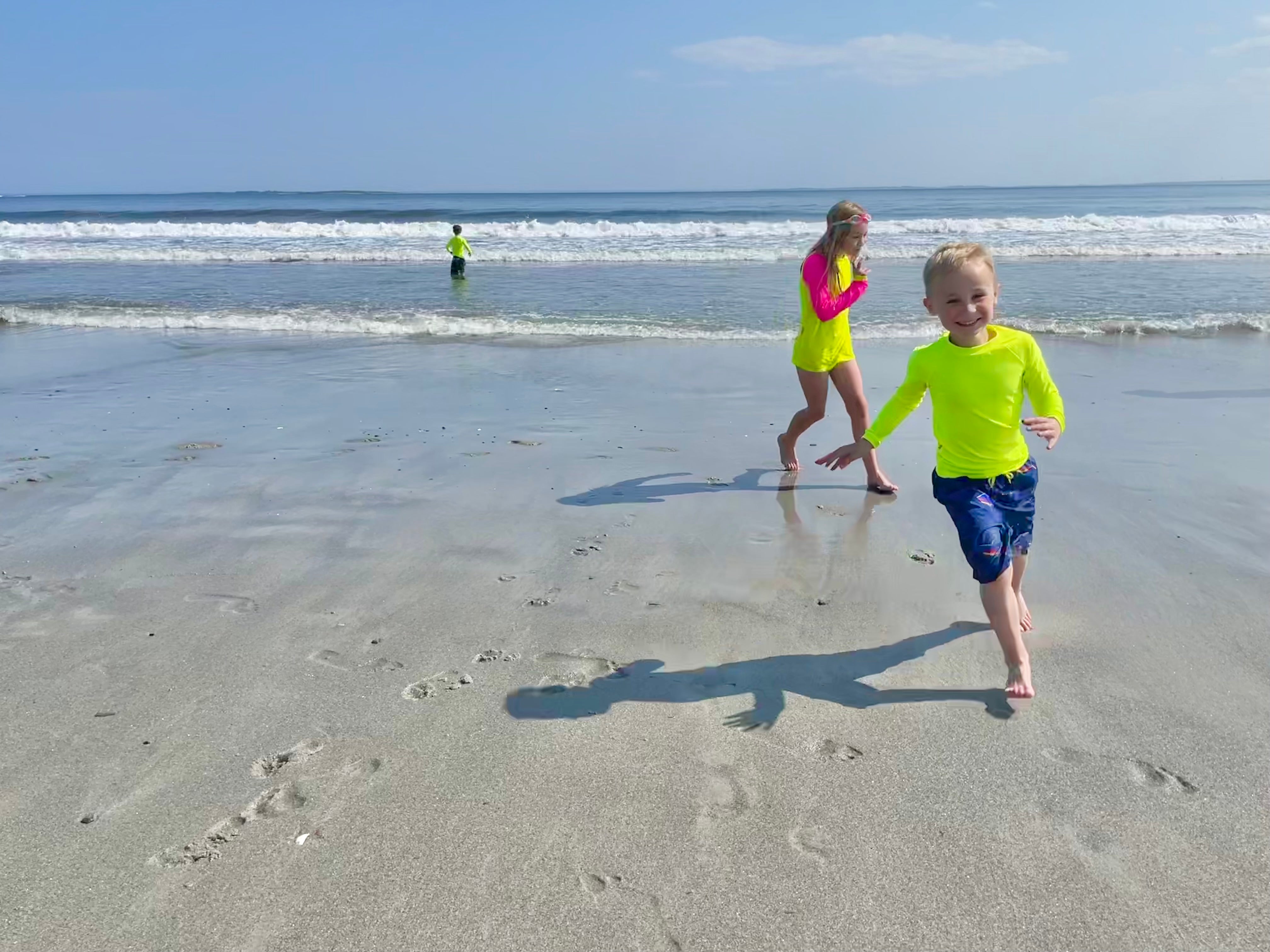 A boy running on the beach in a neon yellow rash guard being chased by his sister also wearing a neon yellow rash guard with neon pink sleeves.