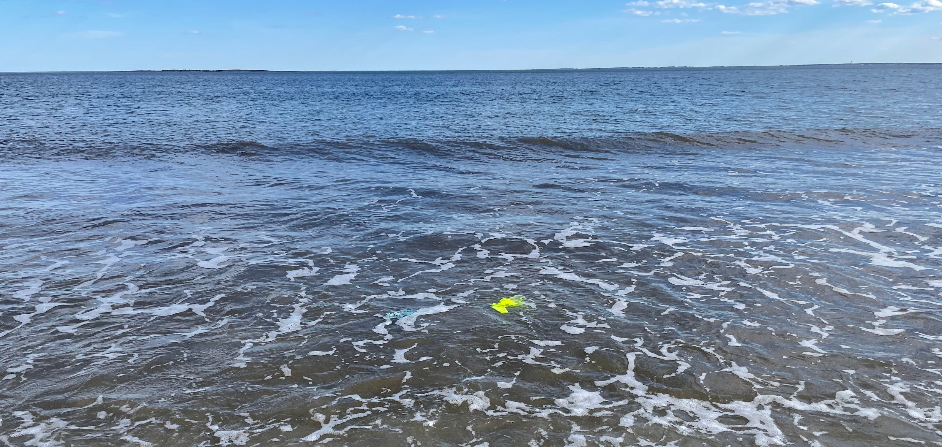 Photo of the ocean just past where the water meets the beach. There is a bright neon swimsuit in the water to right of center and a pale blue, almost invisible one to the left of center. Demonstrates the visibility of neon under water.