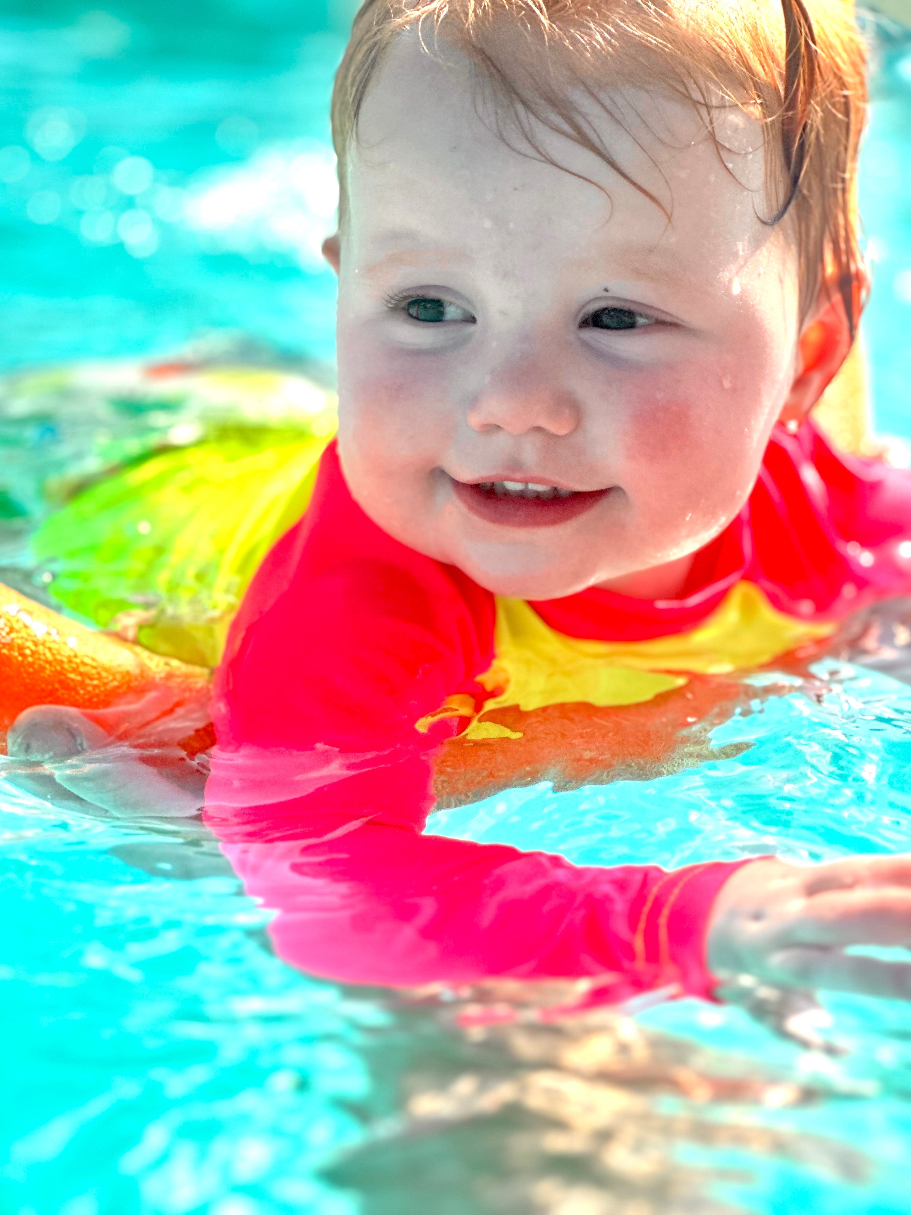 Toddler swimming with parent in a pool outfitted in a bright toddler rash guard with neon pink sleeves and a neon yellow body. It makes her standout from the background. 