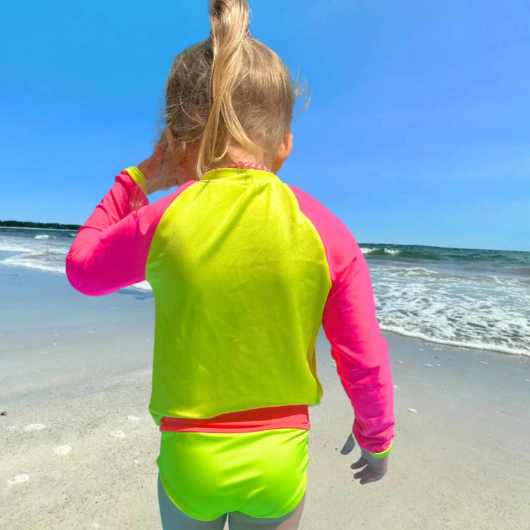A kid standing on the beach back to the camera and facing the water. She is wearing a neon yellow rash guard with neon pink sleeves. She also has on neon swim bottoms from a neon bikini set with a neon pink waistband. She has one hand up on her face and the other by her side. 