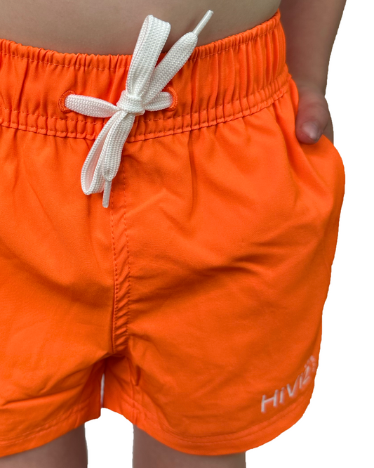 Boy wearing neon orange swim shorts with white fully functional drawstring and hands in his pockets. The neon orange vibrantly stands out against the background. 