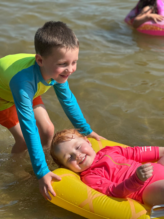 Young girl relaxing in the water wearing a neon pink long sleeve one piece swimsuit with a neon yellow zipper. This high-visibility girls swimsuit is designed for beach days, lake swimming, pool play, and water safety, featuring bright neon colors that help kids stand out in and around the water.