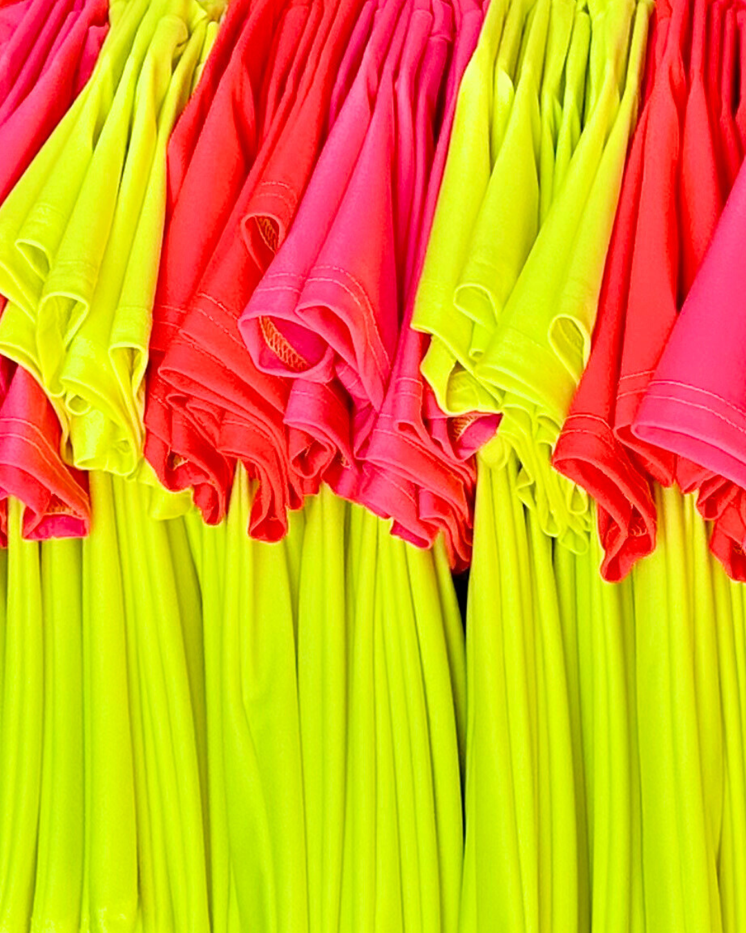 Product image of bright swimwear, short-sleeve neon rash guards, handing with sleeves facing the camera. Neon pink rash guard, neon red rash guard, neon yellow rash guard. 