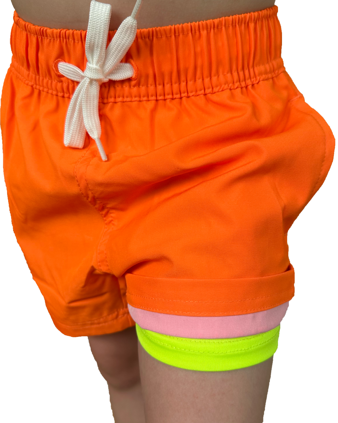 Boy wearing neon orange swim shorts with white fully functional drawstring. The orange swim short shell is rolled up so that you can see the neon yellow boxer brief swim short liner.  The neon orange and neon yellow vibrantly stands out against the background. 