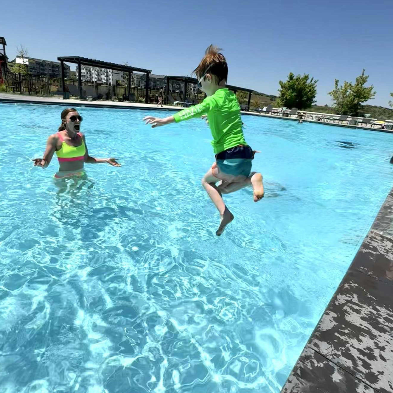 Image of a woman in the pool wearing a neon yellow bikini with her arms out to her sides as a young boy jumps in wearing a boys’ neon green rash guard.  