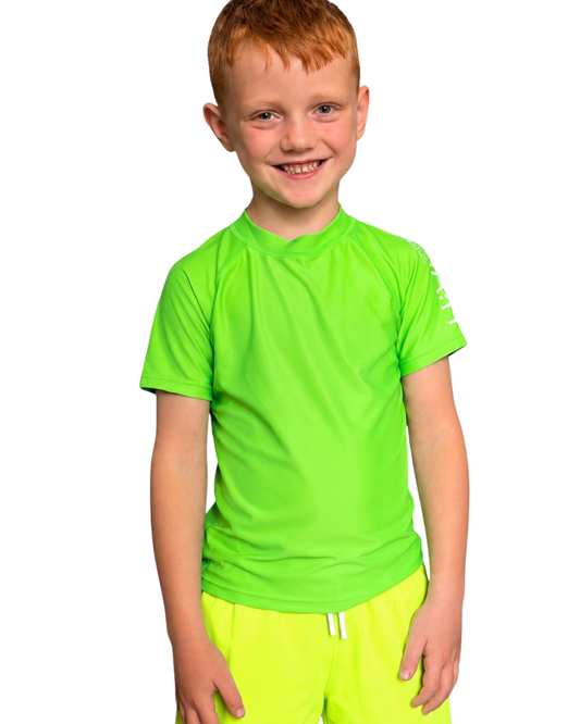 A smiling young boy with red hair wearing a bright neon green hiviz short-sleeve rash guard and matching neon yellow hiviz swim trunks, photographed against a solid white background.