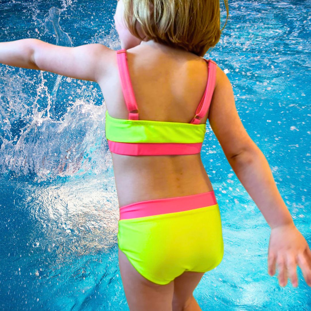 Kid with neon yellow and neon pink bikini swimwear twirling with water background.