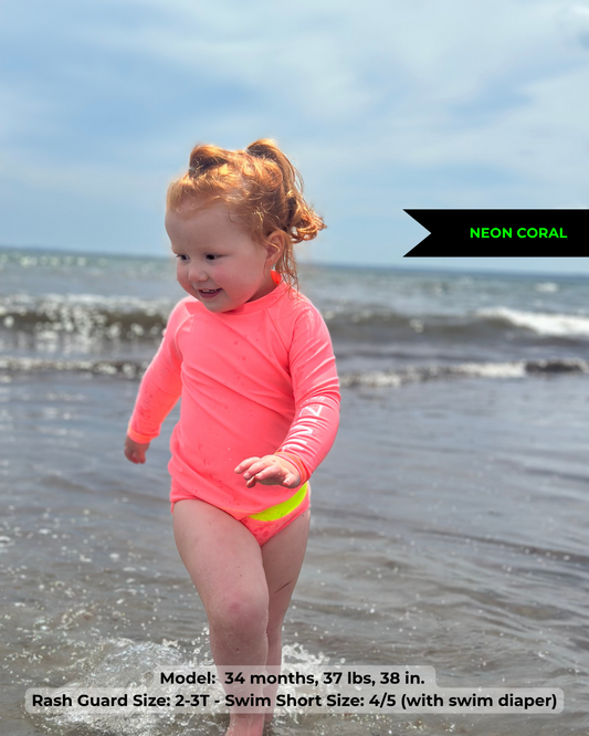 Toddler girl, wearing neon pink coral rash guard and neon coral swim bottoms over cloth swim diaper, runs in the ocean waves of Maine and brightly glows standing out from the back ground. She is incredibly visible in her bright bathing suit. 