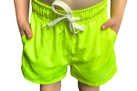 Toddler wearing bright swim shorts, neon yellow swim trunks with his hands in the pockets. 