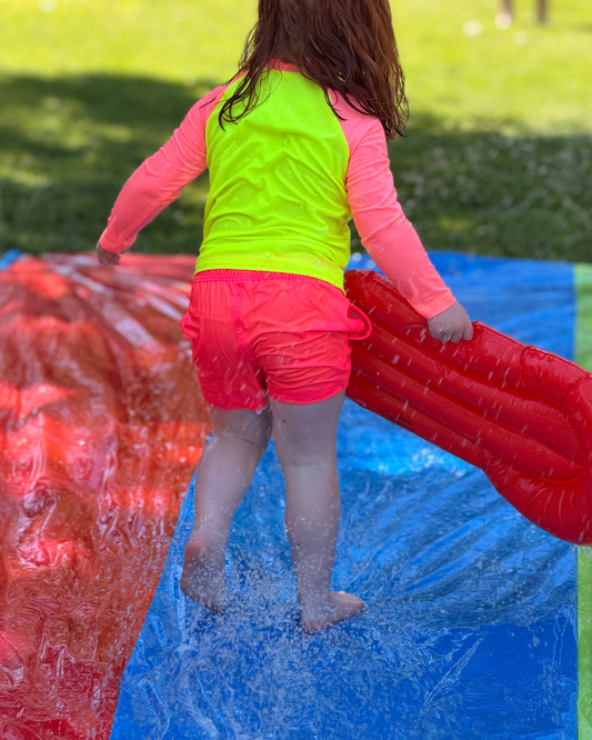 Child in neon pink HiViz Swimwear swim shorts with soft athletic fabric liner enjoying backyard water play. High-visibility kids swimwear helps children stand out at pools, beaches, lakes, camps, and water parks while providing UPF 50+ protection.