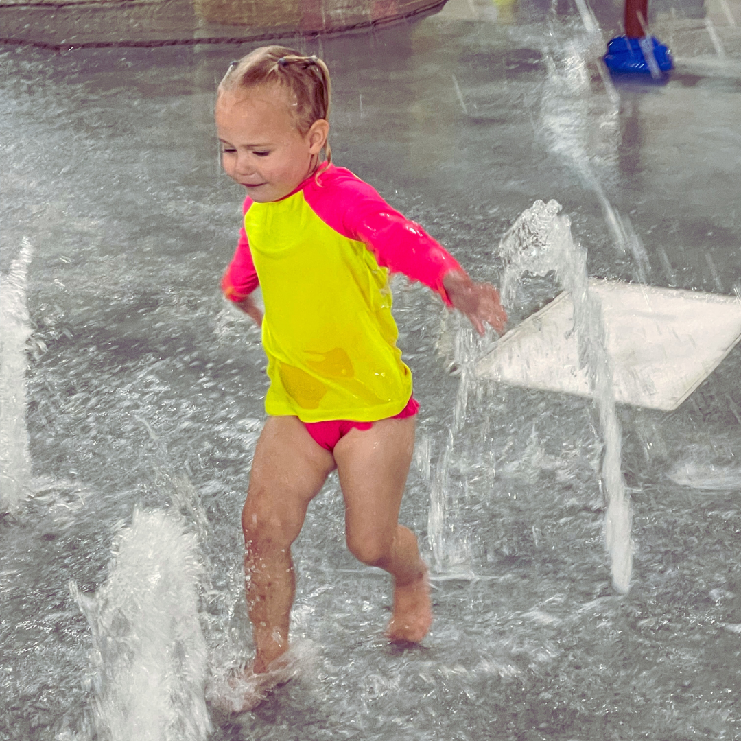Child playing in a water fountain wearing a hiviz neon yellow and neon pink rash guard.