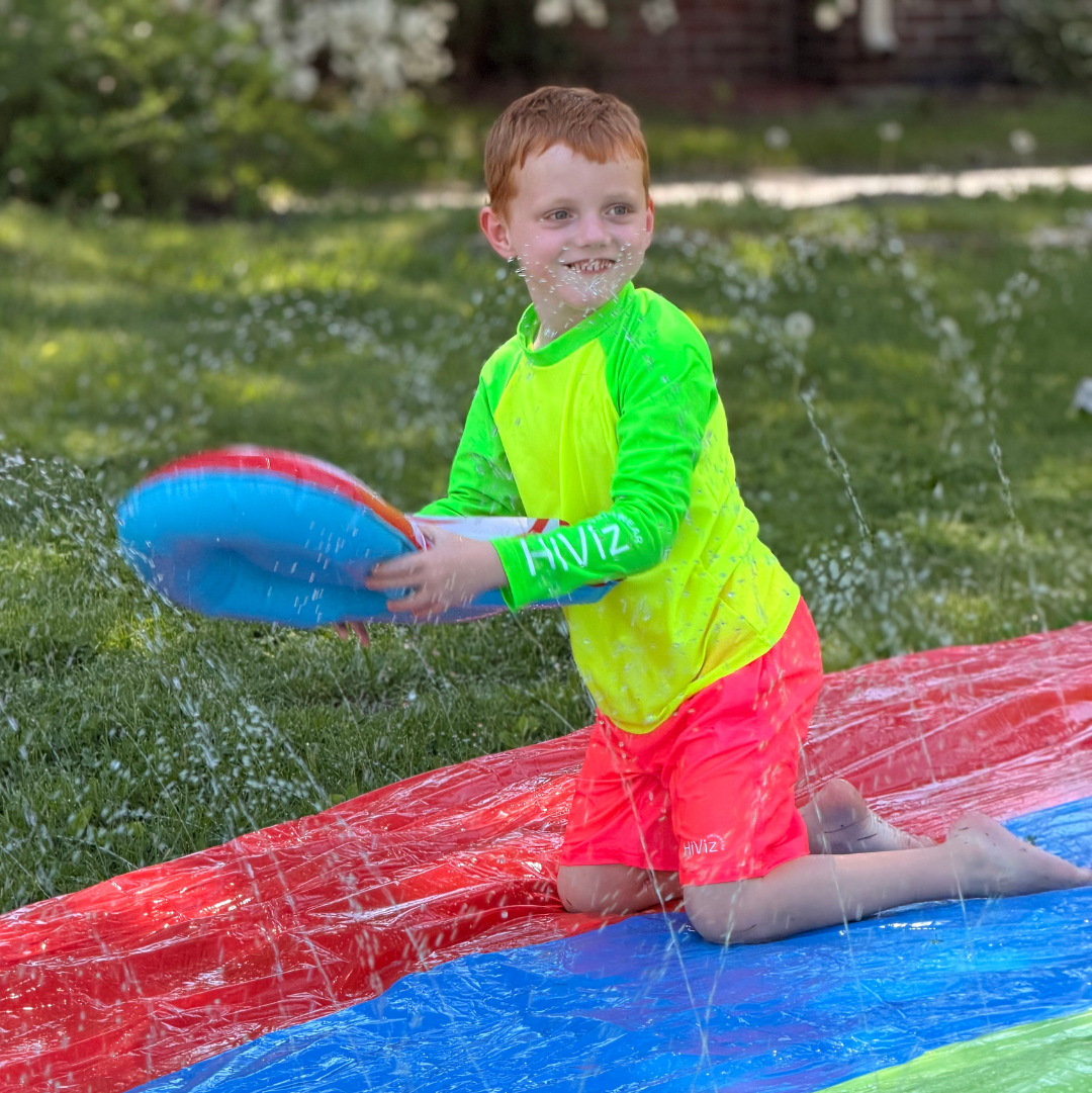 Boy wearing hiviz neon swim shirt and boys neon pink swim trunks on a slip-n-slide smiling and holding a water toy in his hands.