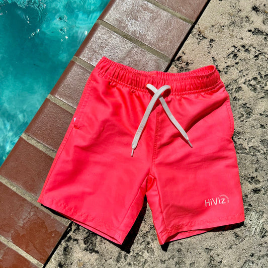 HiViz boys neon swim trunks laying on a concrete patio next to a pool. These boys neon swim trunks are electric neon pink.