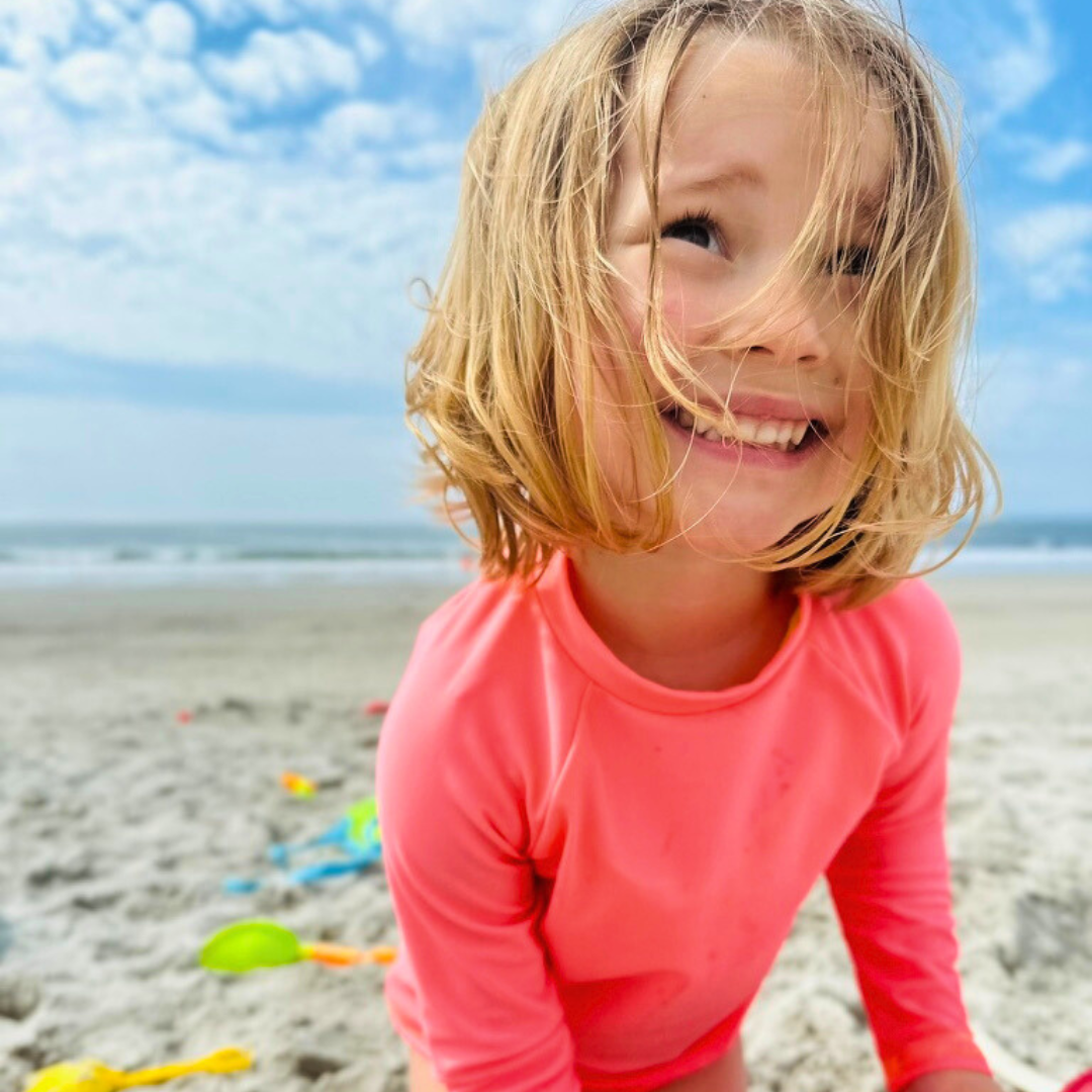Young girl at the beach loking up to the right smiling. She is wearing bright swimwear, a neon rash guard.