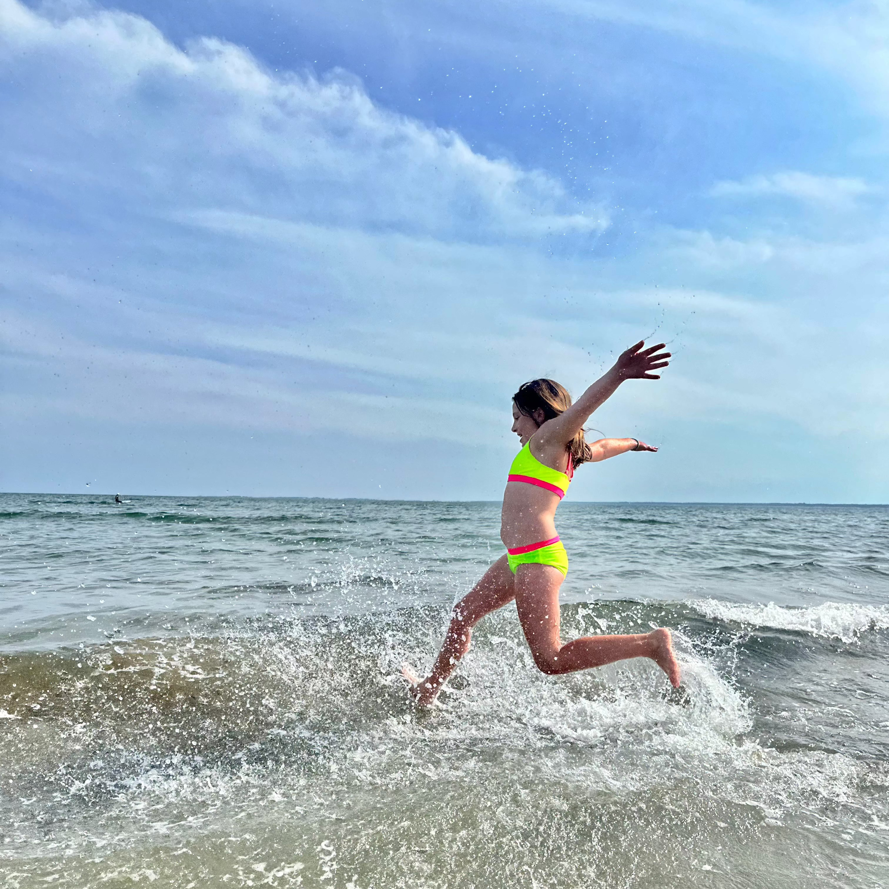 Tween girl jumping waves wearing a neon yellow and neon hot pink bikini from the bright swimwear line from HiViz. Her bright swimsuit stands out from the dull background. Neon is the best swimsuit color for visibility because of this.