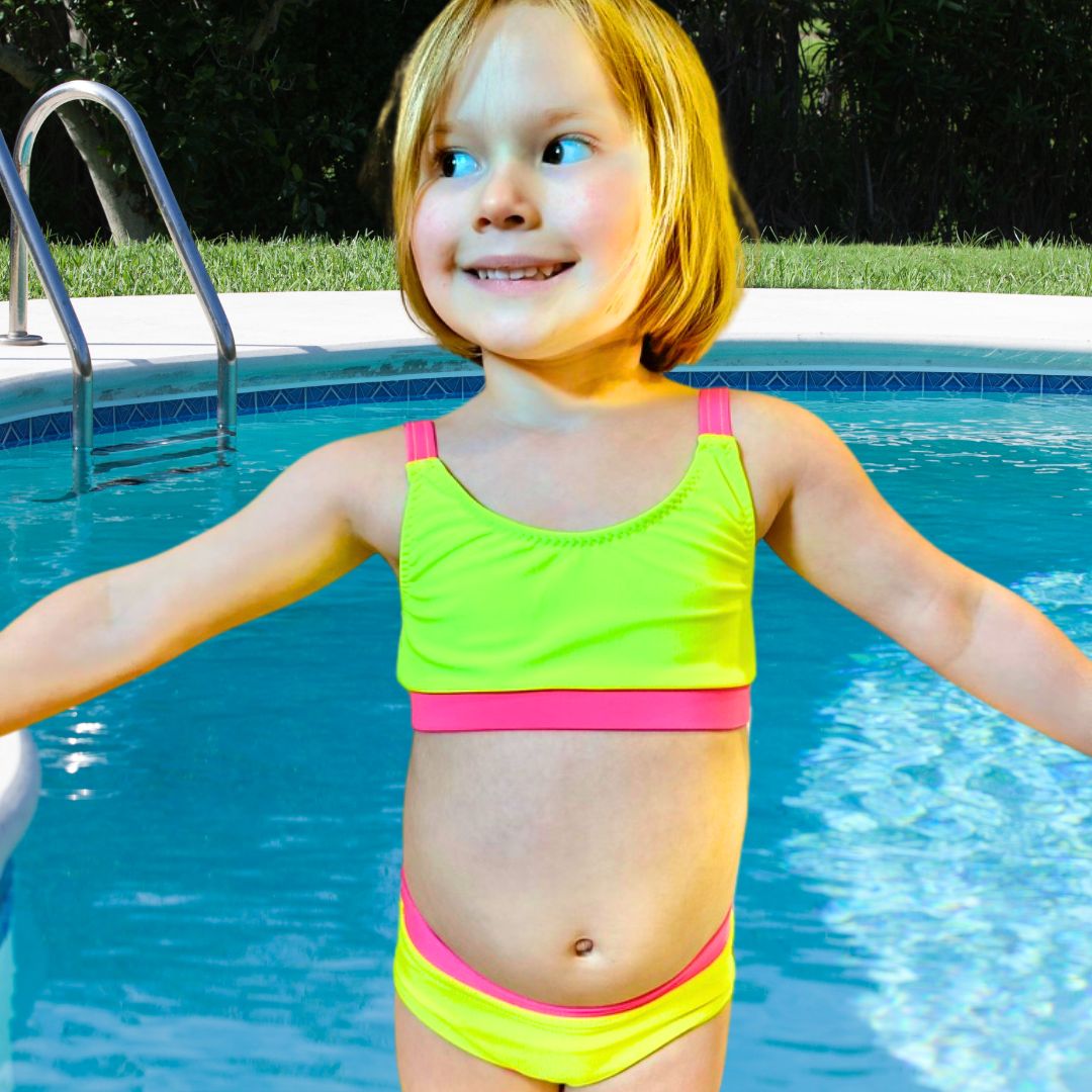 Girl in bright swimwear, a yellow neon bikini, smiling with her arms out to the side and a blue pool backdrop. 