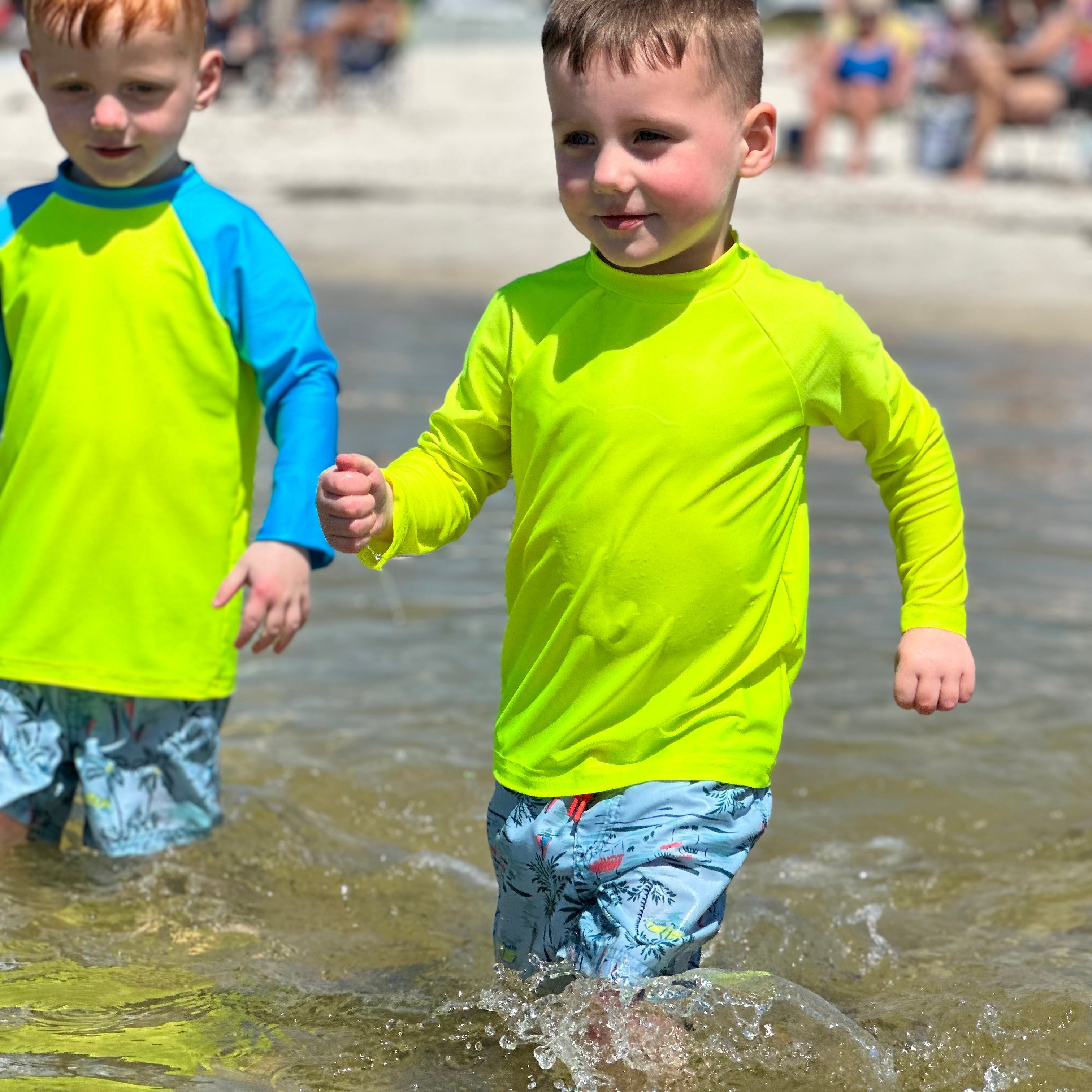 A kid running into the ocean wearing a neon yellow rash guard with another pre-school aged boy next to him wearing a neon yellow and blue sleeved rash guard. The bright swimwear stands out against the dark water.