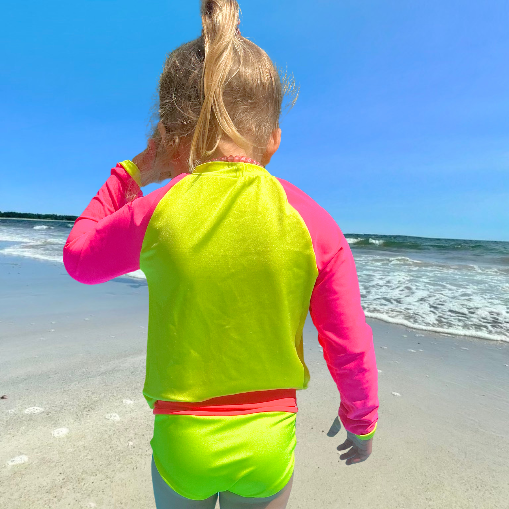 A kid standing on the beach back to the camera and facing the water. She is wearing a neon yellow rash guard with neon pink sleeves. She also has on neon swim bottoms from a neon bikini set with a neon pink waistband. She has one hand up on her face and the other by her side.  Her bright swimwear makes her stand out from the dull background.