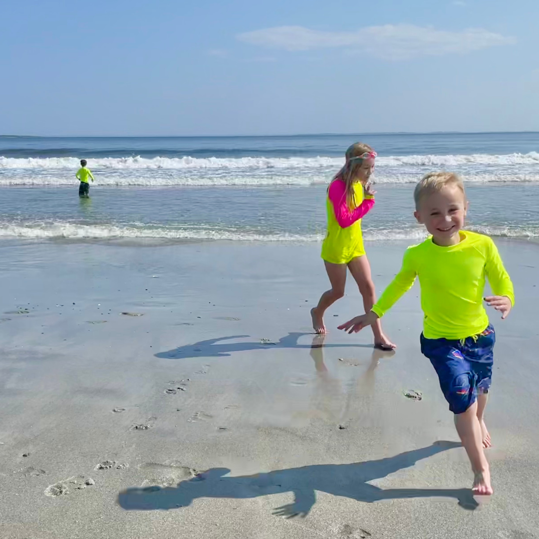 Three kids on the beach with neon yellow rash guards on from the true neon, bright swimwear company, HiViz Swimwear. These kids stand out in their bright swimwear agains the dull backdrop.