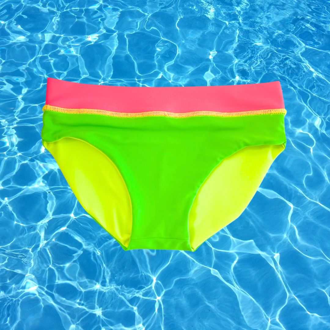 Product image of neon green reversible swim bottom from HiViz's bright swimwear. The bright swimsuit bottoms are on a backdrop of rippling blue pool water.