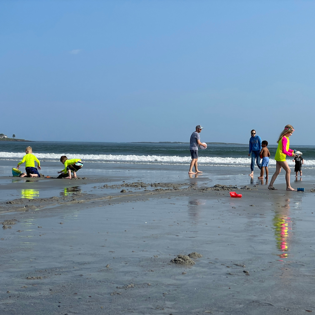 Three kids on the beach in neon swimwear, neon yellow rash guards, contrasting to four other people who are not in neon and blend in. These kids in their bright swimwear stand out of the dull background while the people not in bright swimwear blend in.