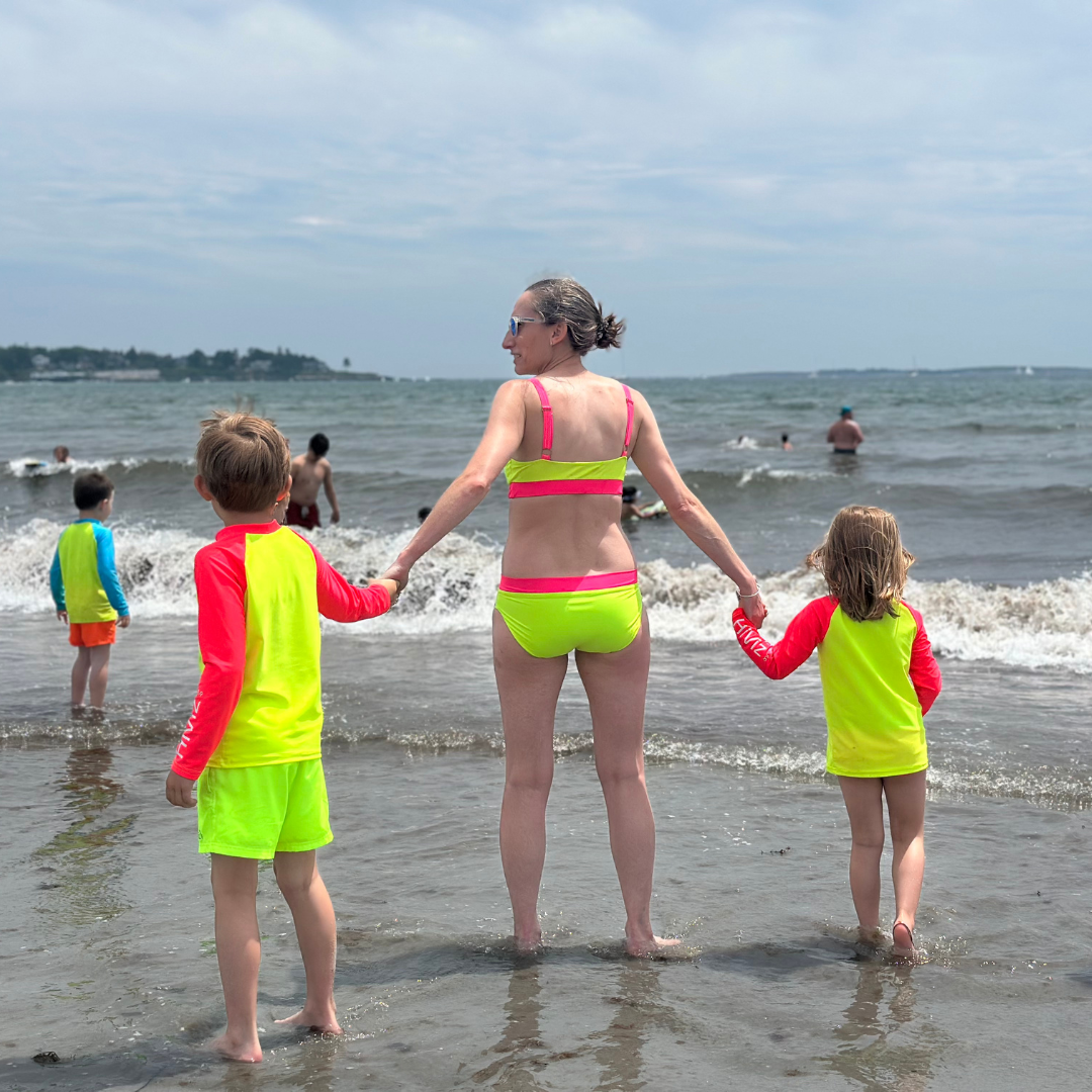 Woman and two children in bright neon swimwear standing on a beach with ocean waves.