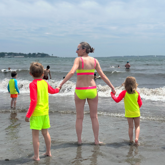 Woman and two children in bright neon swimwear standing on a beach with ocean waves.