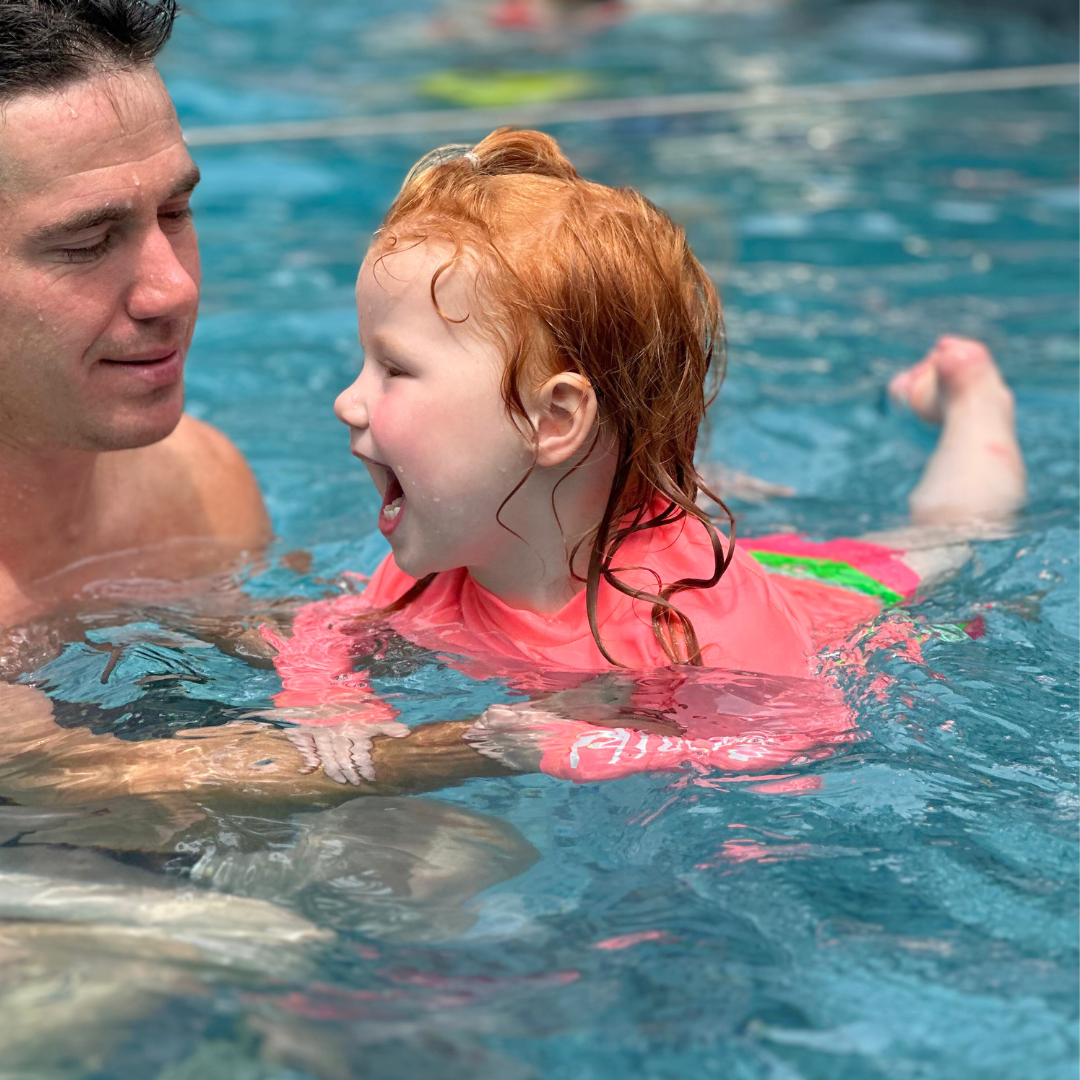 Girl floating on her belly in the pool laughing with her dad. She's wearing a neon pink rash guard.