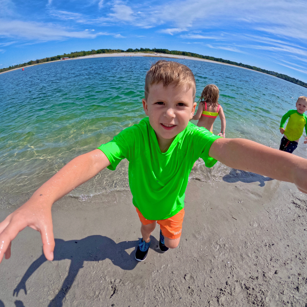 Child in a hiviz neon green rash guard and hiviz neon orange swim shorts standing on a beach with clear blue water and sky.