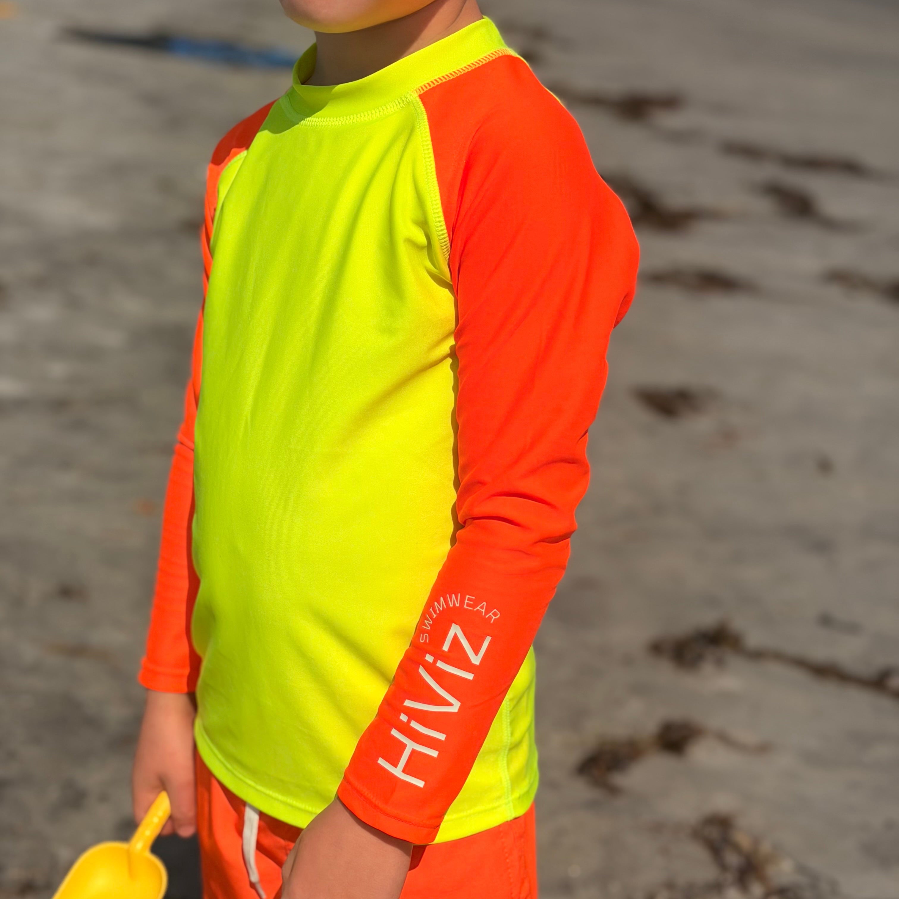 A child wearing a bright neon yellow and orange long-sleeve rash guard and matching orange swim shorts on a sandy beach, holding a yellow shovel.
