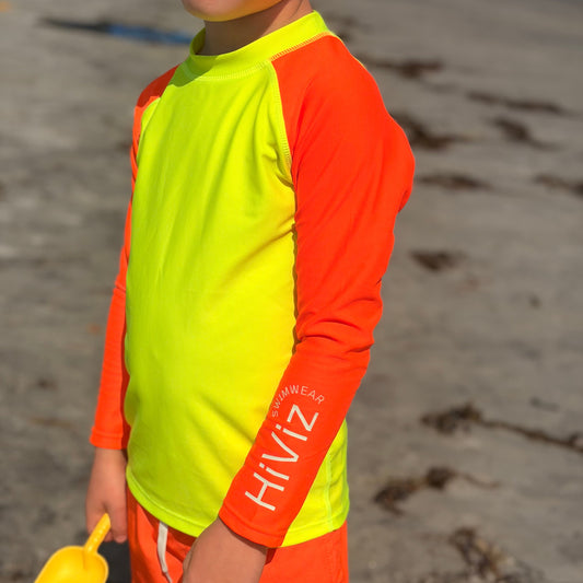 A child wearing a bright neon yellow and orange long-sleeve rash guard and matching orange swim shorts on a sandy beach, holding a yellow shovel.