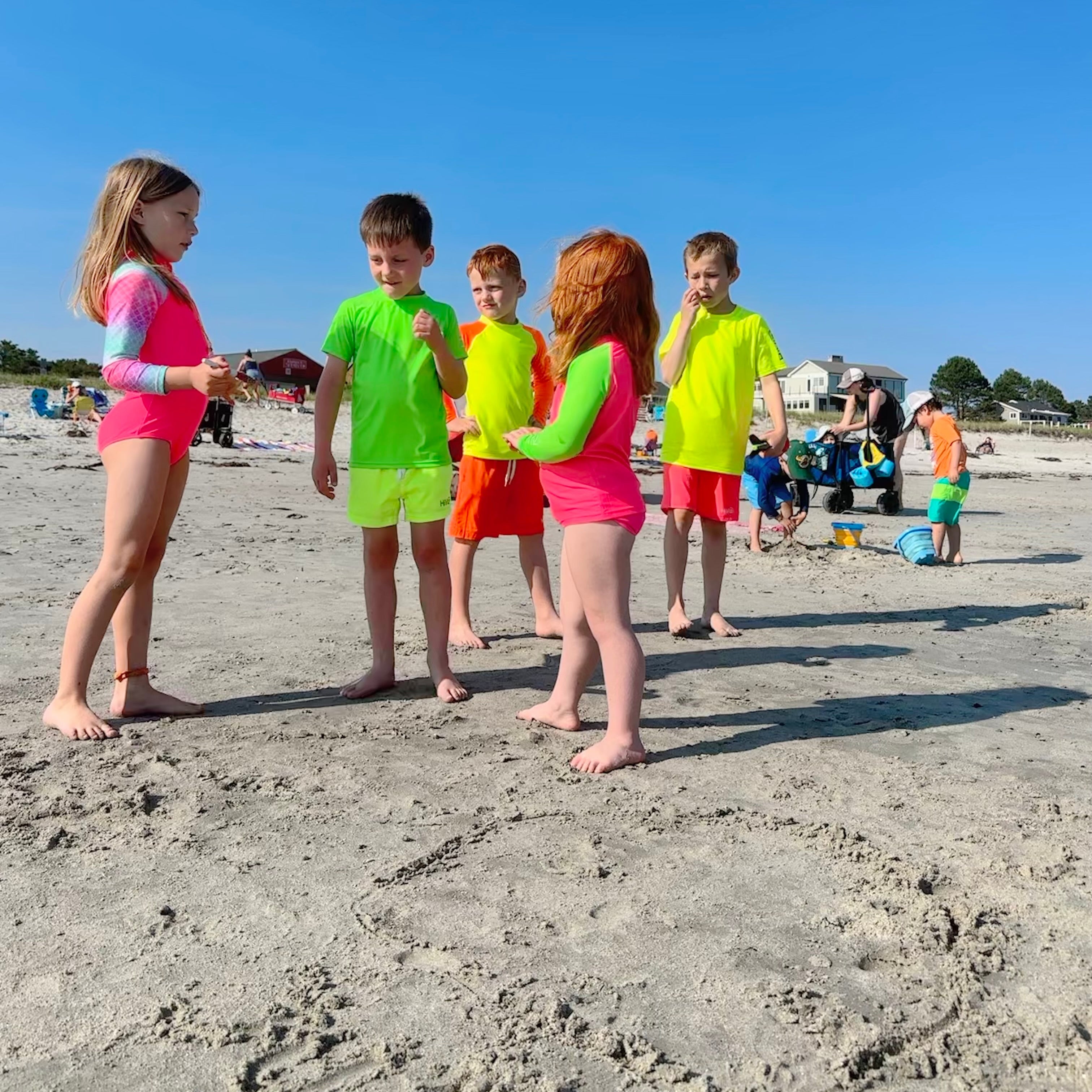 Five children wearing bright neon swim shirts and neon swim trunks standing on a sandy beach under a clear blue sky.