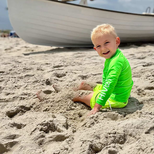 Child in hiviz neon green swimsuit rash guard and hiviz neon yellow swim trunks playing in the sand with a boat in the background