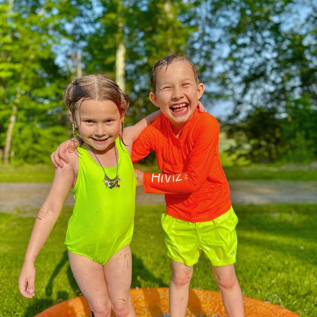 Two children in hiviz neon swimwear standing on a slip and slide with trees in the background