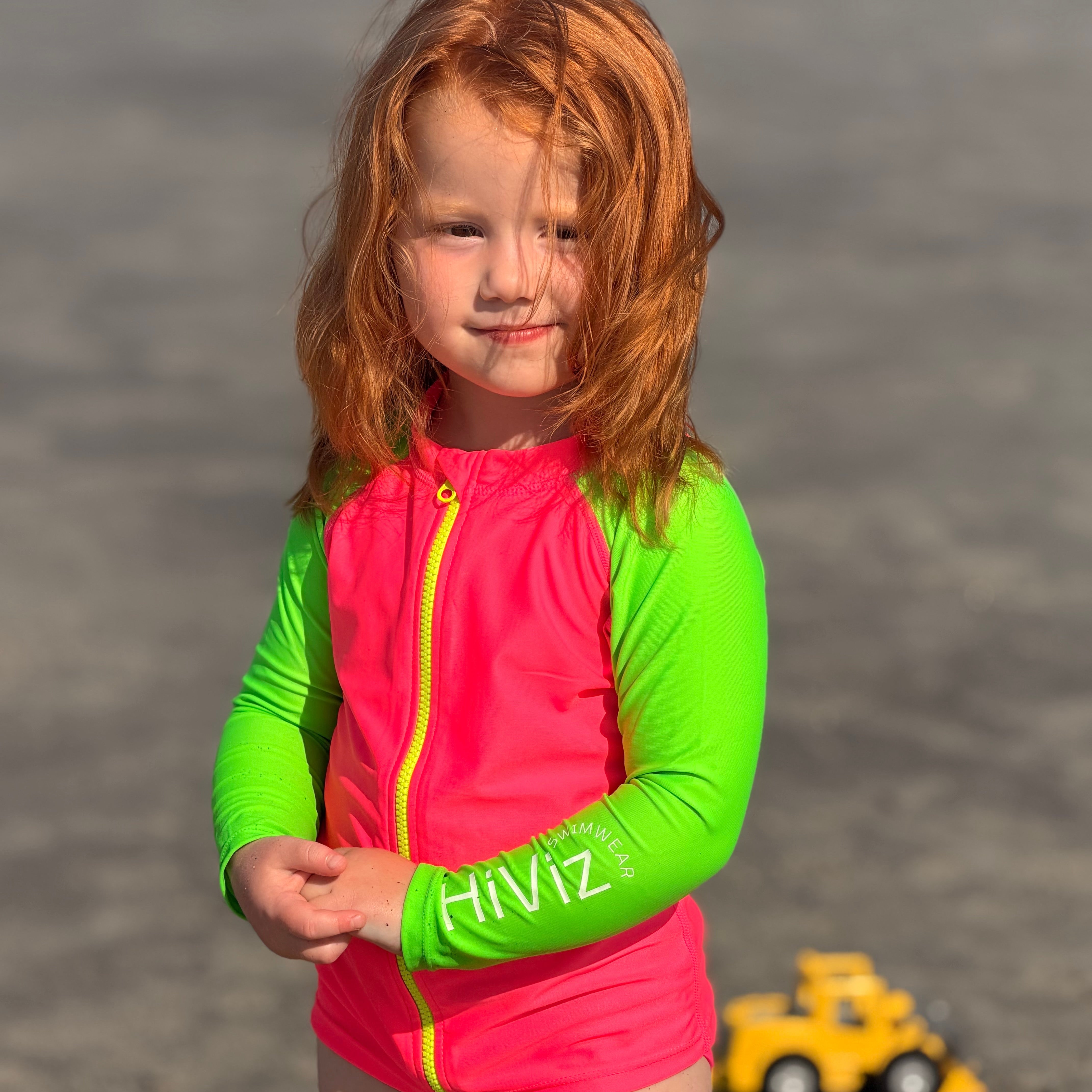 Child wearing a hi-viz neon pink and neon green zip-up rash guard with 'HIVIZ' on sleeves, standing on a beach with toys in the background.