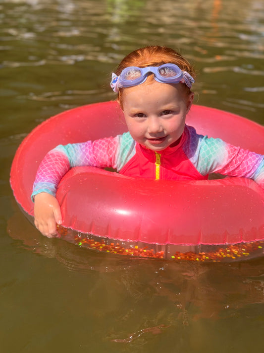 Child wearing kids’ high-visibility neon swimwear and goggles in a bright pink swim float—hi-vis safe swimwear that stands out in the water and helps make children easier to spot while swimming.