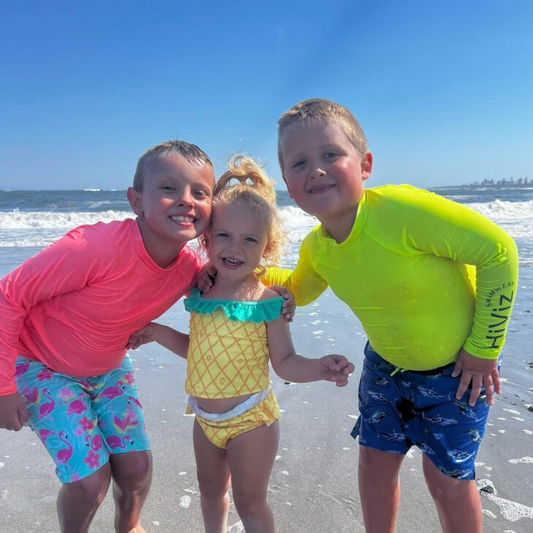 Three children two wearing neon rash guards on a beach with ocean waves in the background