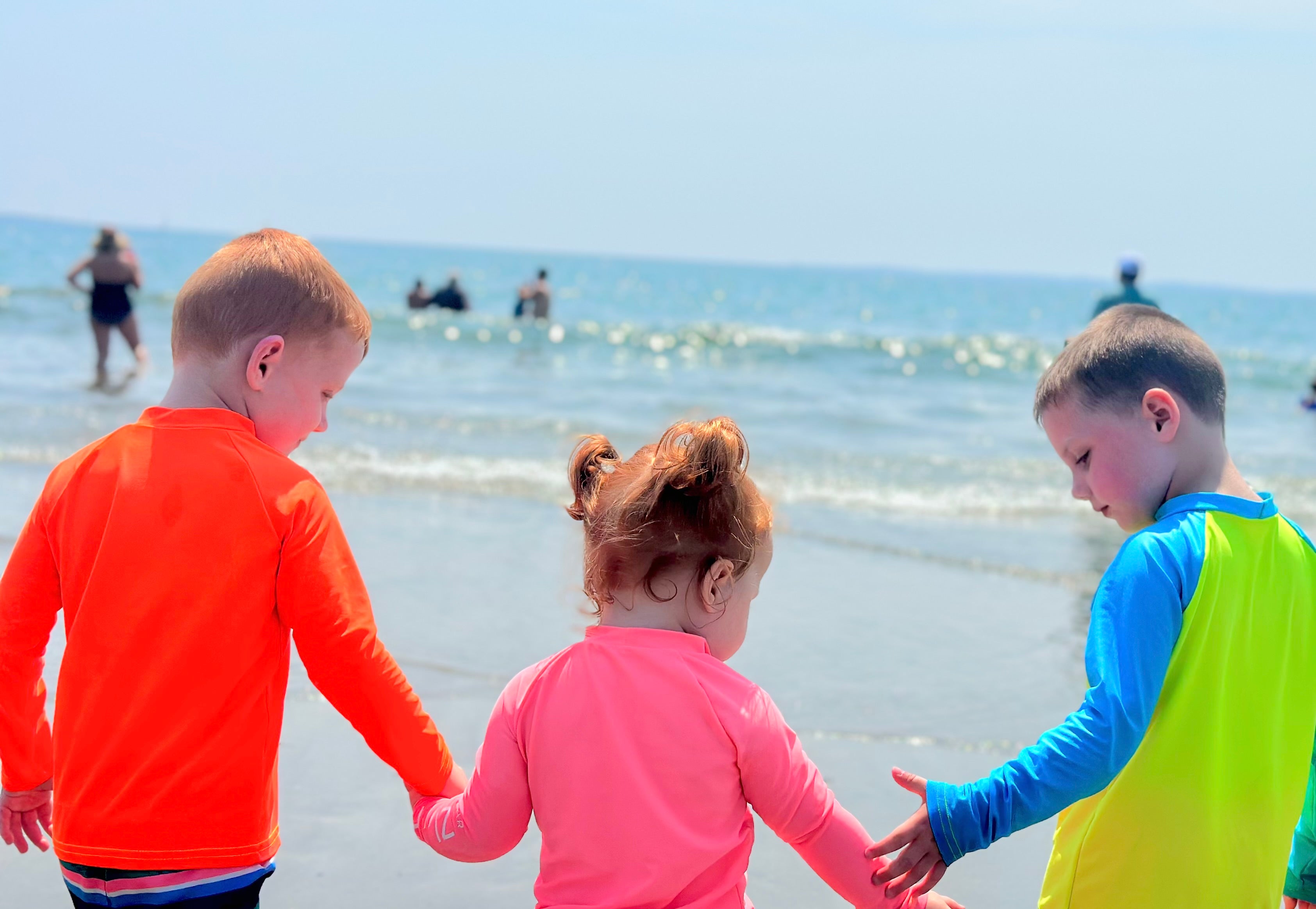 kids neon orange rash guard, kids neon coral rash guard and kids neon yellow rash guard, kids hand in hand walking toward the ocean at the beach.