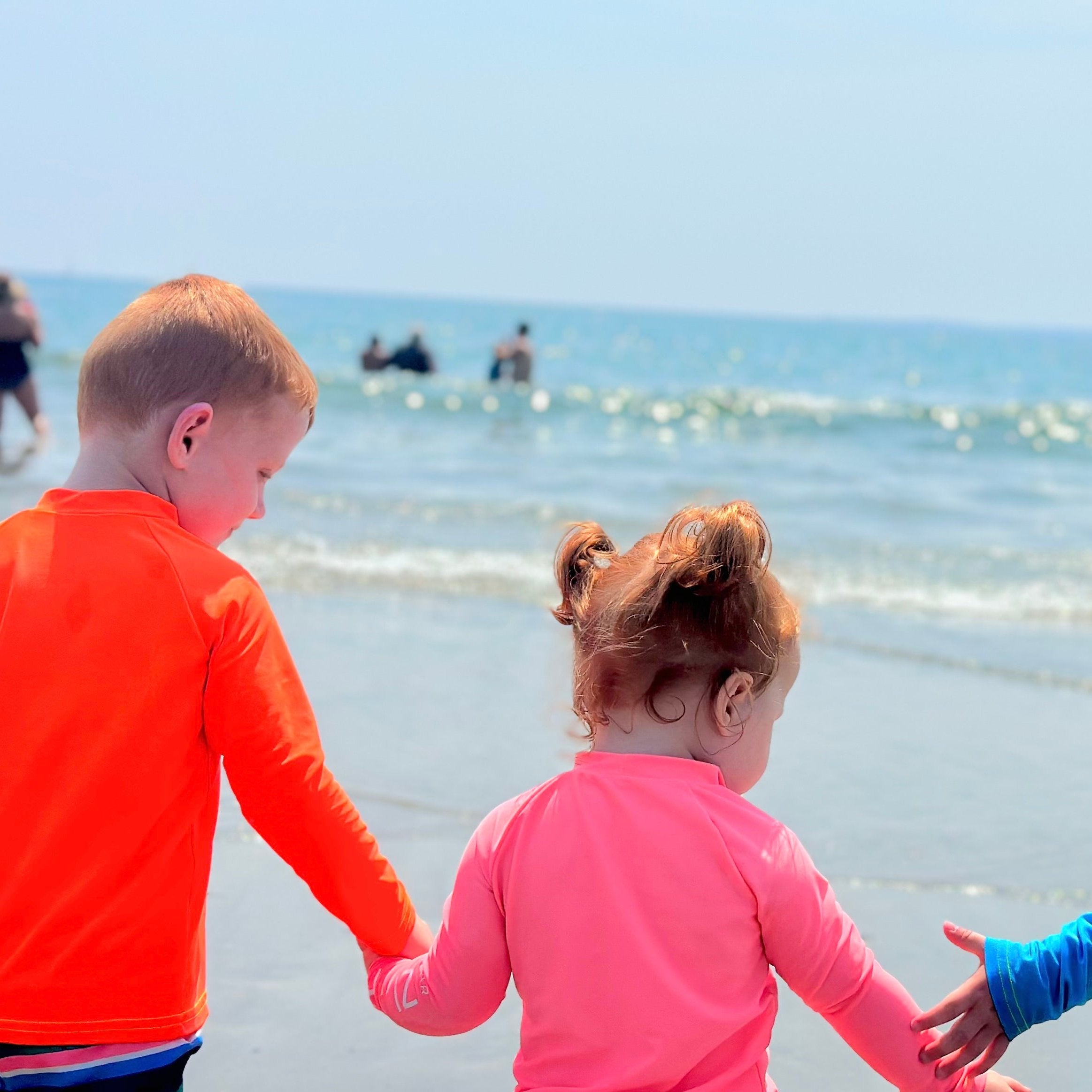 kids neon orange rash guard, kids neon coral rash guard and kids neon yellow rash guard, kids hand in hand walking toward the ocean at the beach.