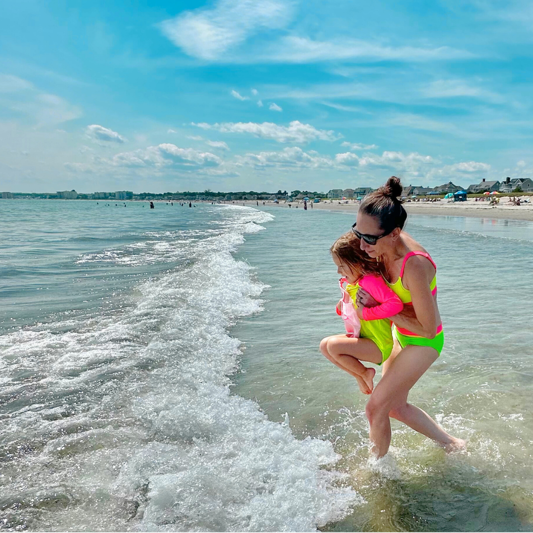 Mom wearing women's bright swimsuit neon bikini with neon yellow top with neon pink accents holding young girl wearing hi vis girls bright swimsuit neon long sleeve rash guard with neon yellow body and neon pink sleeves for sun protection and neon yellow bright swimsuit neon bottoms.