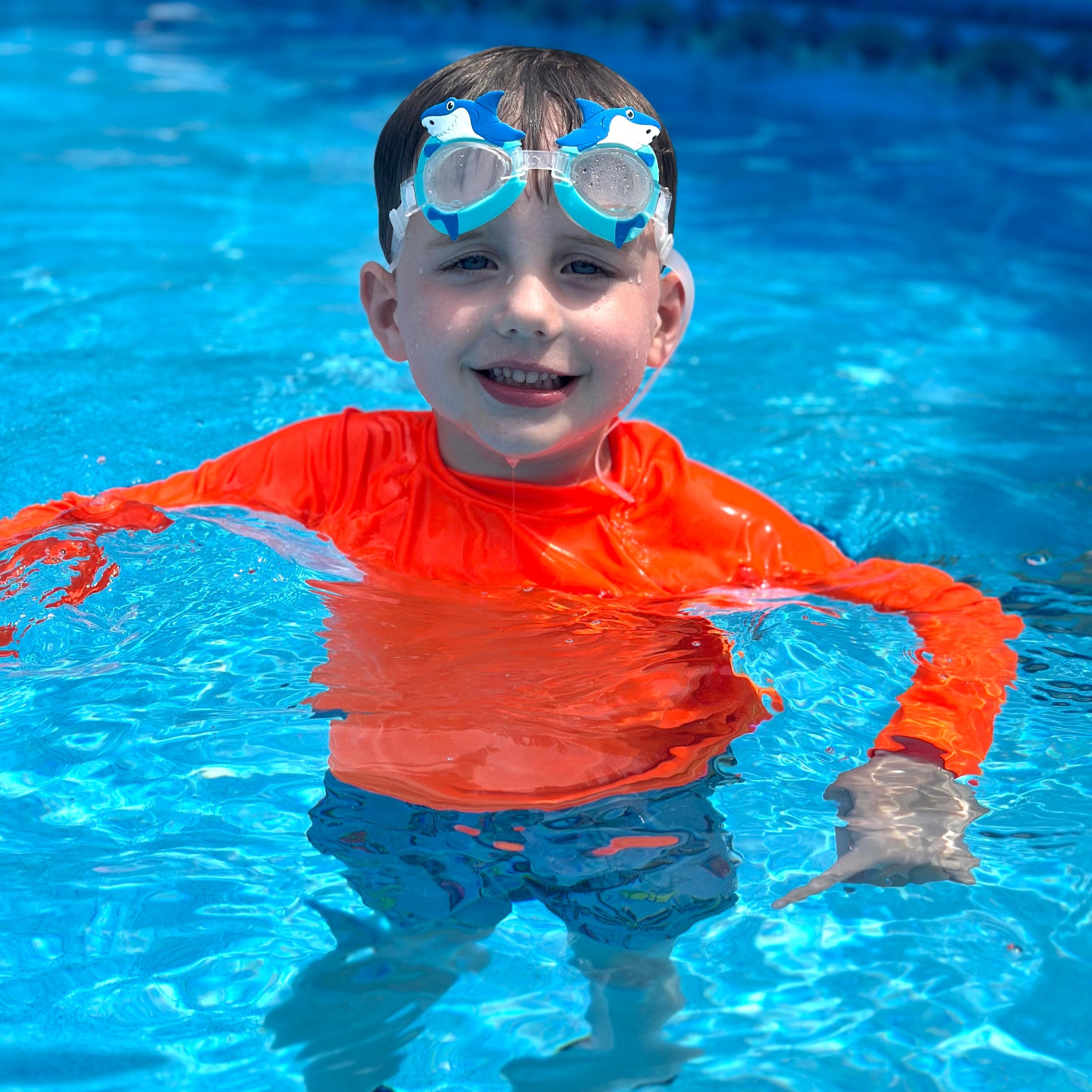 Young boy in a pool up to his armpits in the water. He is smiling, wearing bright swimwear, a neon orange rash guard. The neon rash guard is glowing above and below water. 