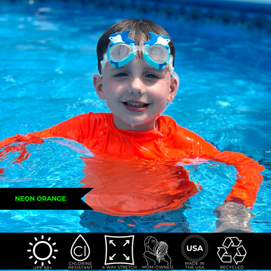 Young boy in a pool up to his armpits in the water. He is smiling, wearing bright swimwear, a neon orange rash guard. The neon rash guard is glowing above and below water. 