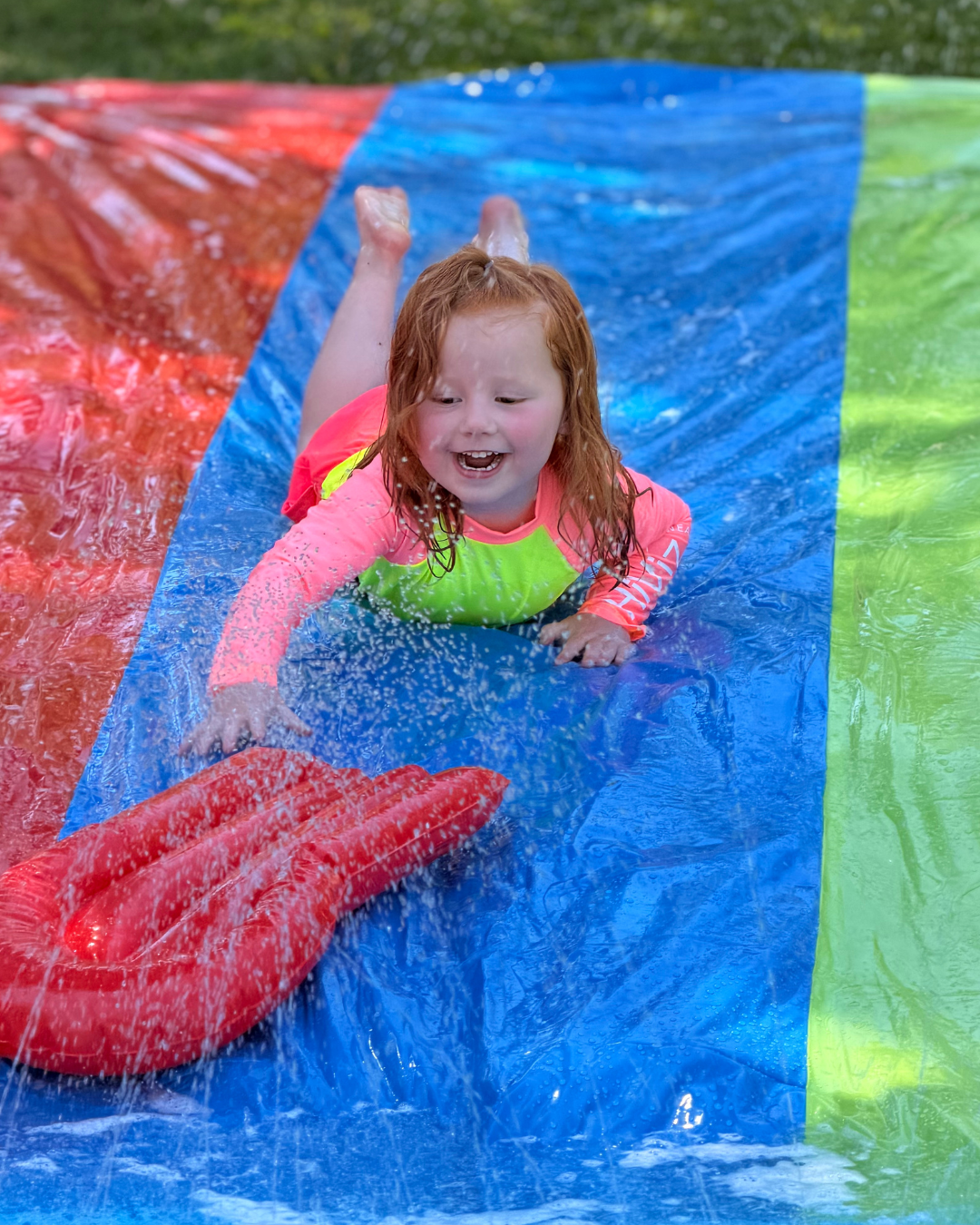 Girl wearing bright neon HiViz Swimwear rash guard and swim shorts playing on a slip-and-slide during summer. High-visibility kids swimwear with UPF 50+ sun protection, designed to make children easier to spot in and around water at pools, beaches, lakes, camps, and water parks.
