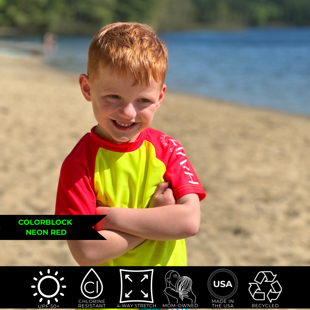 Photo of a young readheaded boy smiling with his arms crossed wearing a neon yellow and neon red rash guard from HiViz Swimwear. 