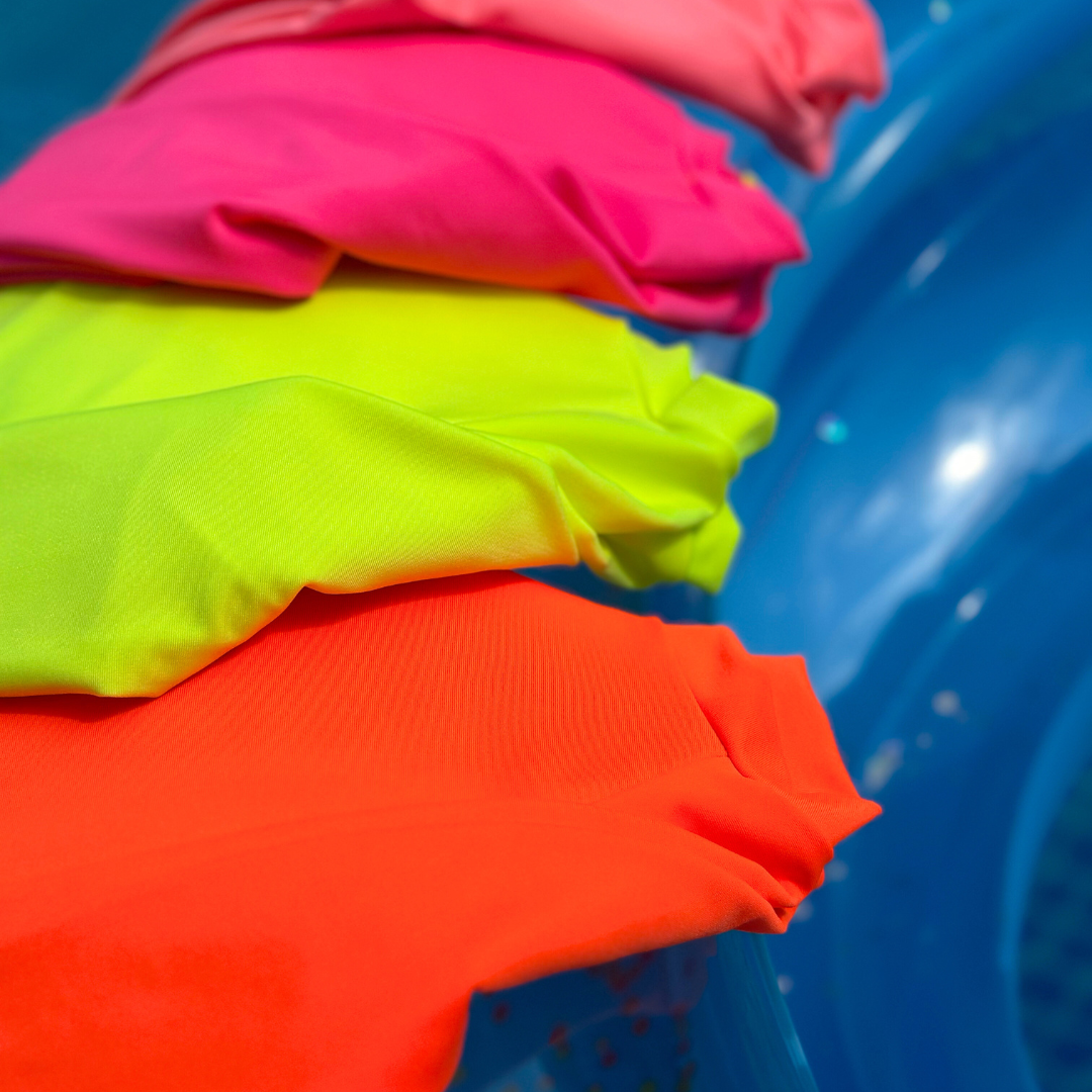 Bright swimwear from HiViz swimwear displaying neon rash guards floating on a pool float. Neon orange rash guard, neon yellow rash guard, neon pink rash guard, and a neon coral rash guard. These bright rash guards are on the left of the picture going from bottom to top.