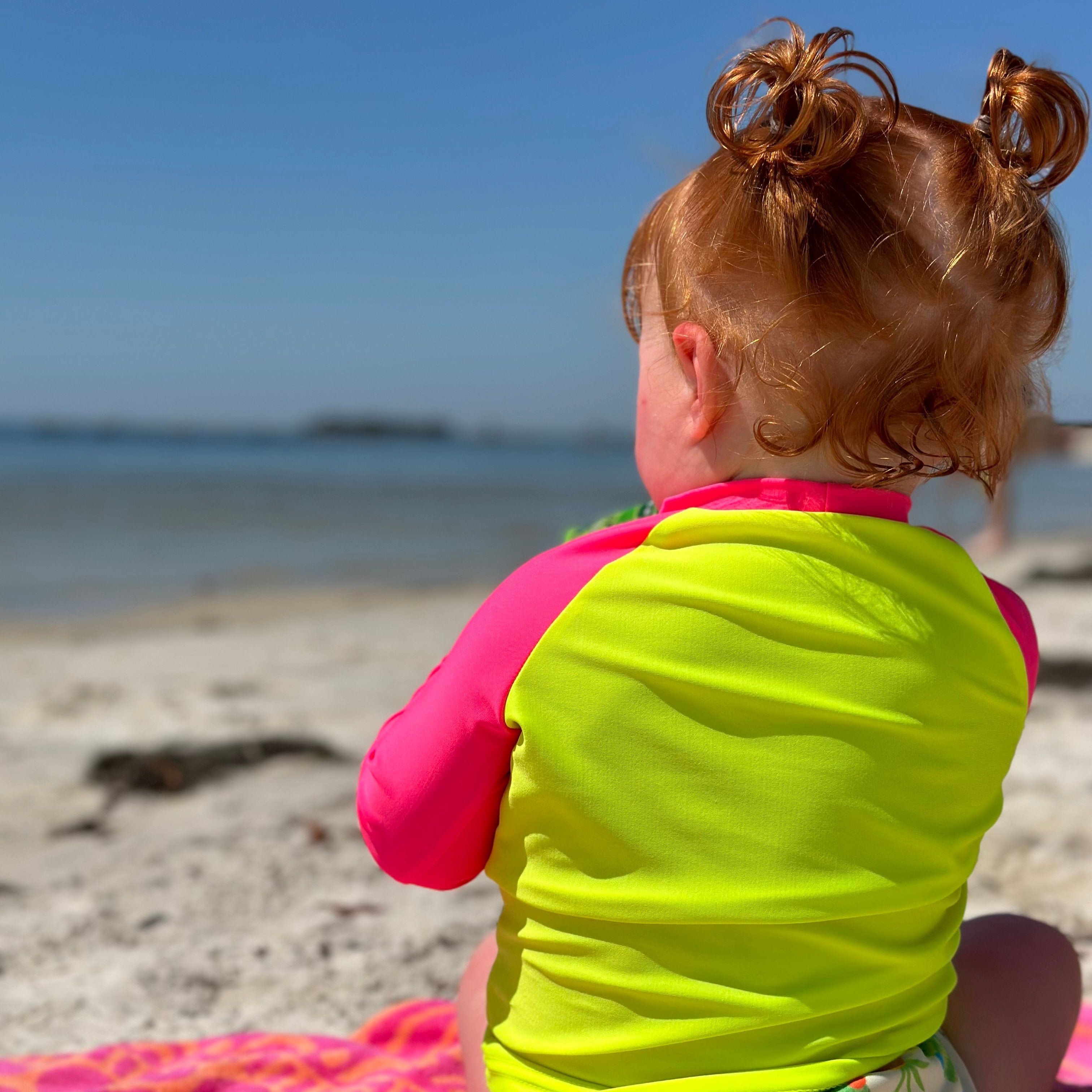 Baby girl sitting on towel on beach sand looking out at ocean wearing a neon yellow rash guard with neon pink sleeves; the bright swimwear stands out against the dull backdrop. 