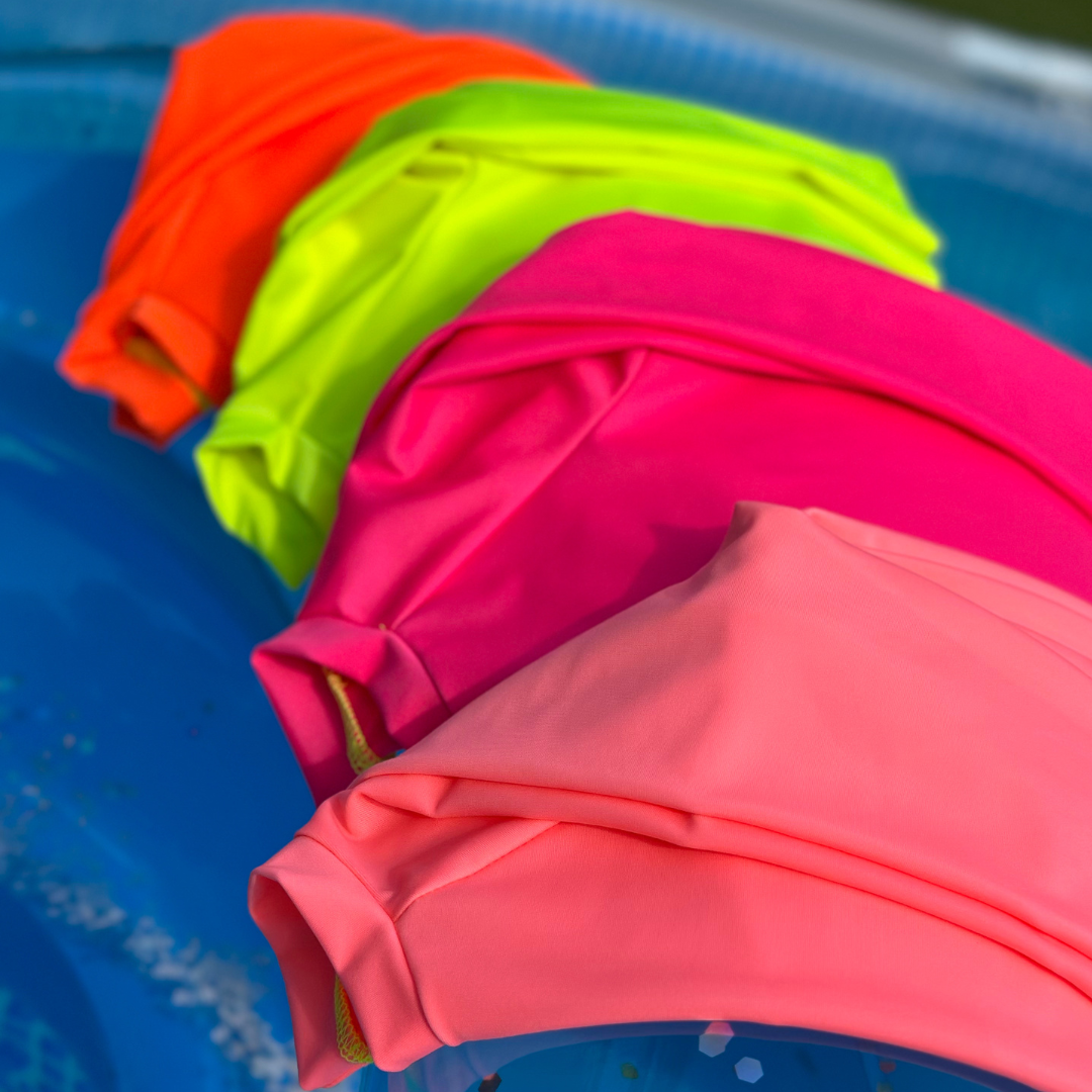 Neon Swimwear, neon coral rash guard, neon pink rash guard, neon yellow rash guard, and neon orange rash guard, sitting on a blue tube floating in a blue pool.