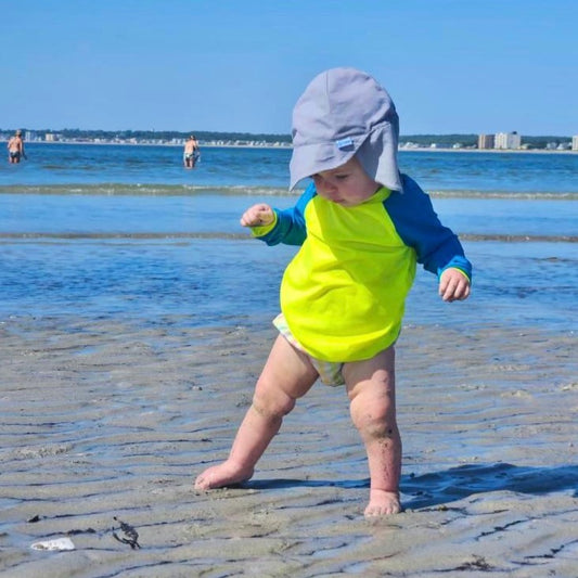 Baby Standing on the Beach in a HiViz Neon Rash Guard for Babies. Neon Yellow Rash Guard With Blue Sleeves.